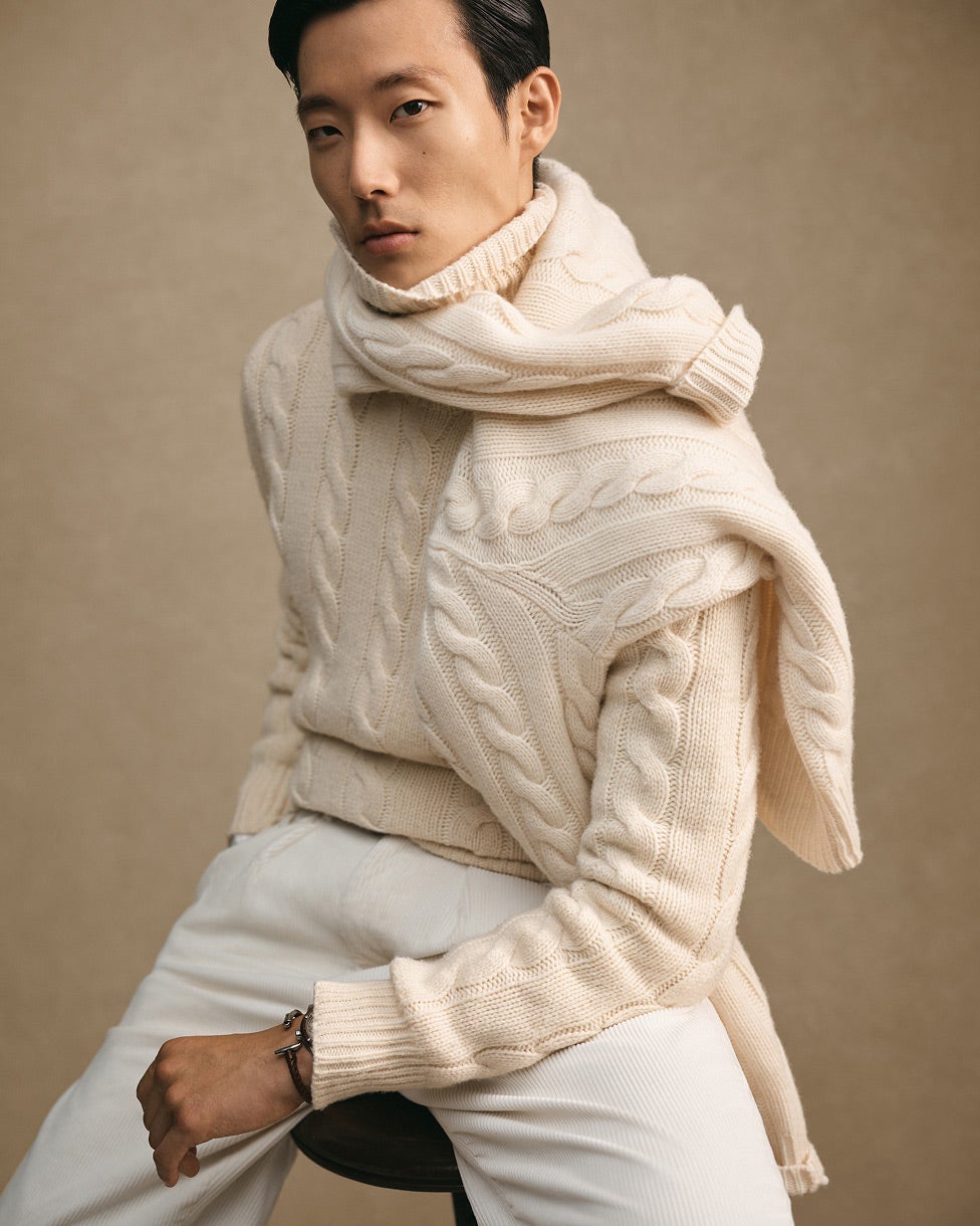 Sitzender Mann in cremefarbenem Pullover mit Zopfmuster, mit passendem Schal und weißer Hose.