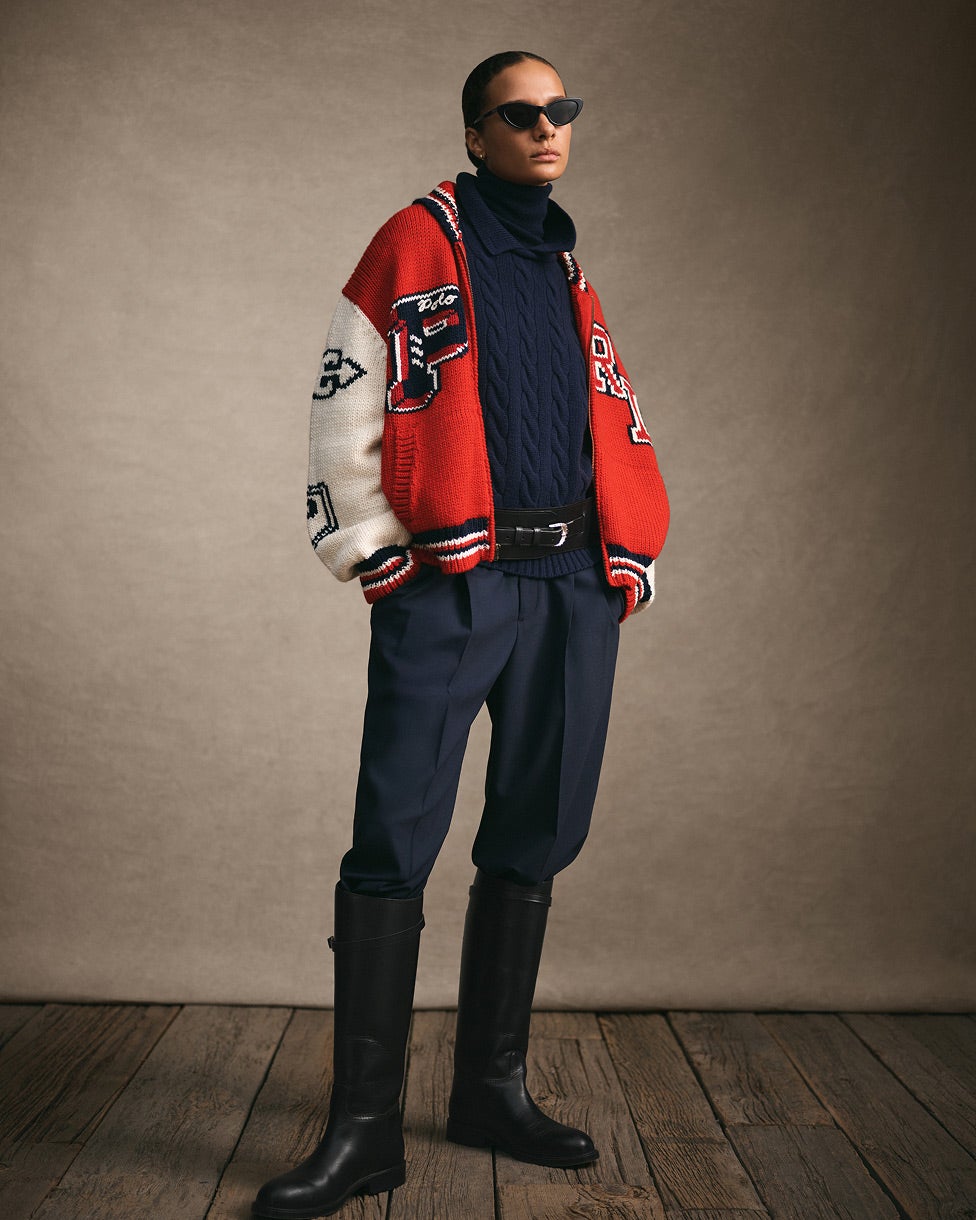 Stehende Frau in rot-weißer College-Jacke mit marineblauem Pullover und marineblauer Hose sowie hohen schwarzen Stiefeln.