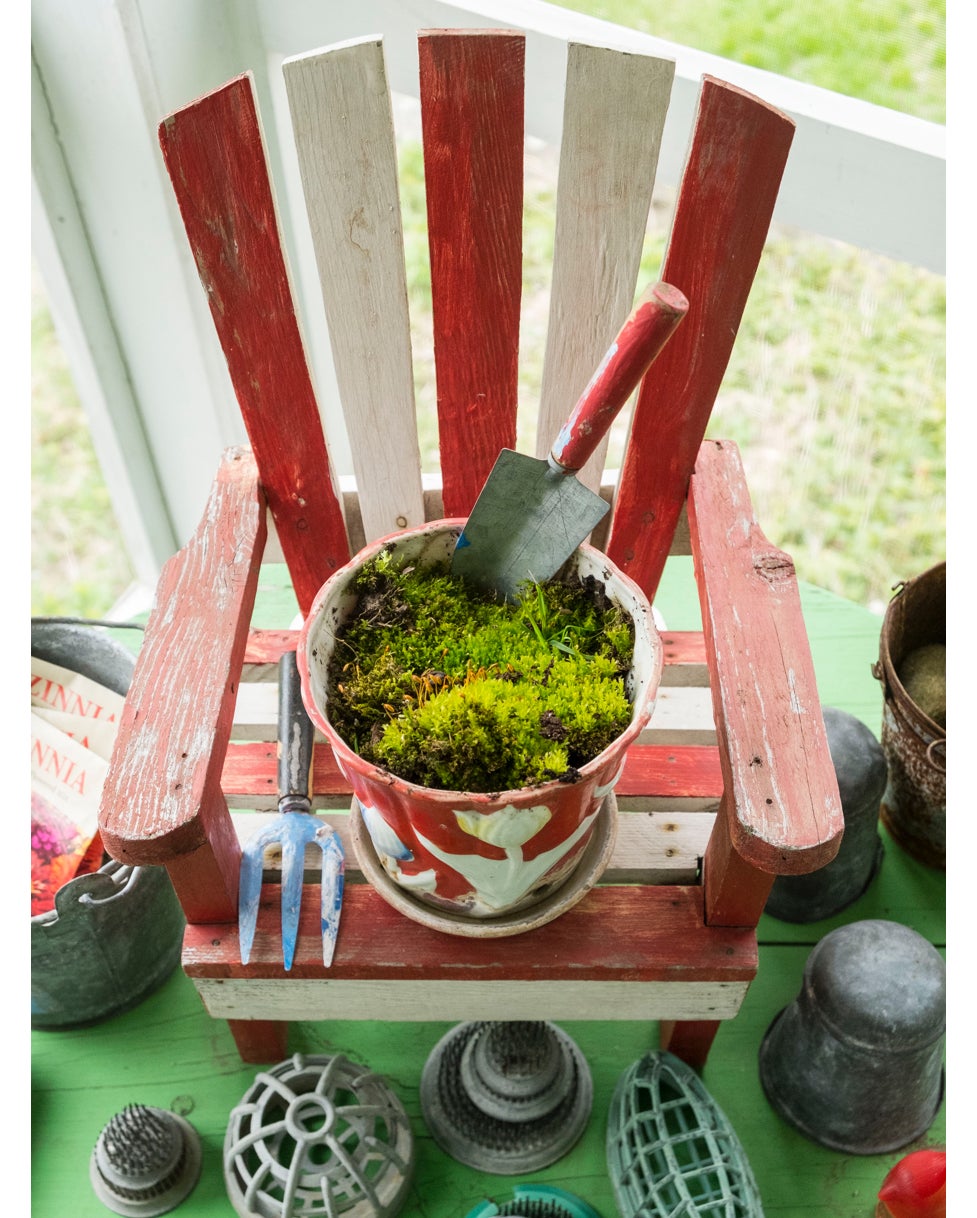 Ein geheimer Garten aus Moos in einem Vintage-Blumentopf schmiegt sich an einen Adirondack-Stuhl in Puppengröße, umgeben von einer Sammlung an Blumenarrangements und Fröschen