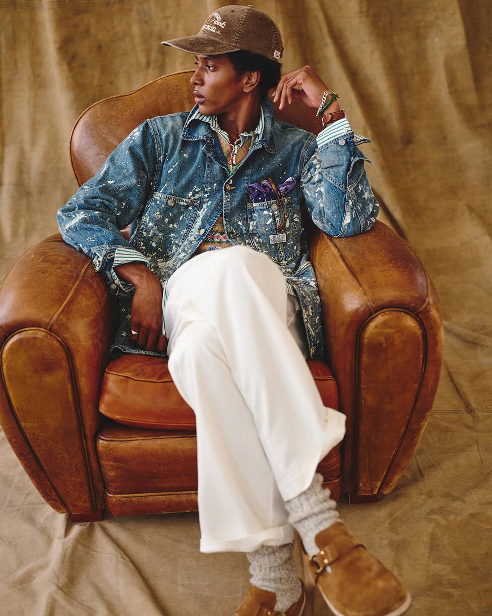 Man seated, wearing a denim jacket, patterned shirt, white trousers, and a cap.