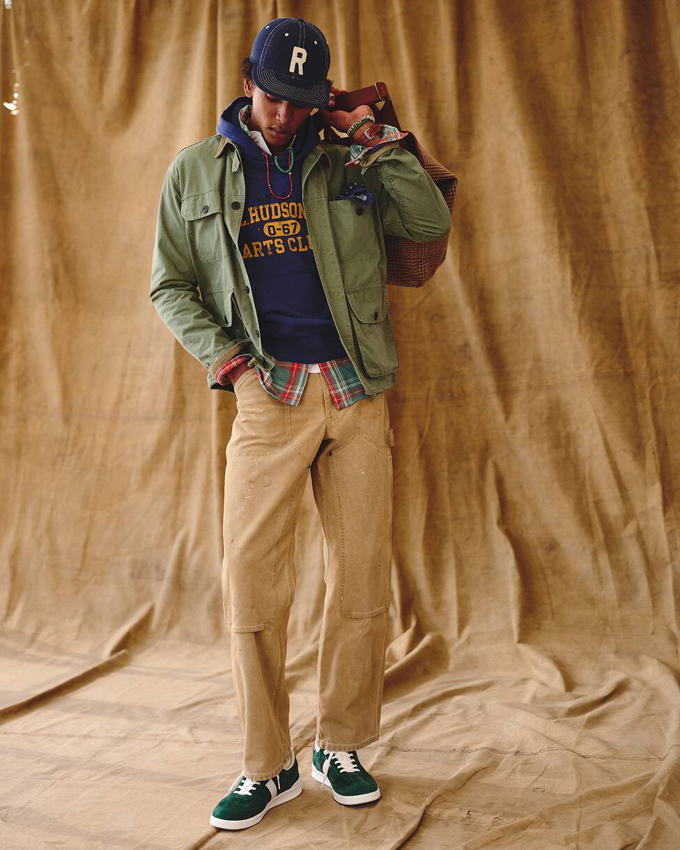 Man wearing a utility jacket layered over a hoodie and checked shirt, with beige trousers and trainers and a cap.