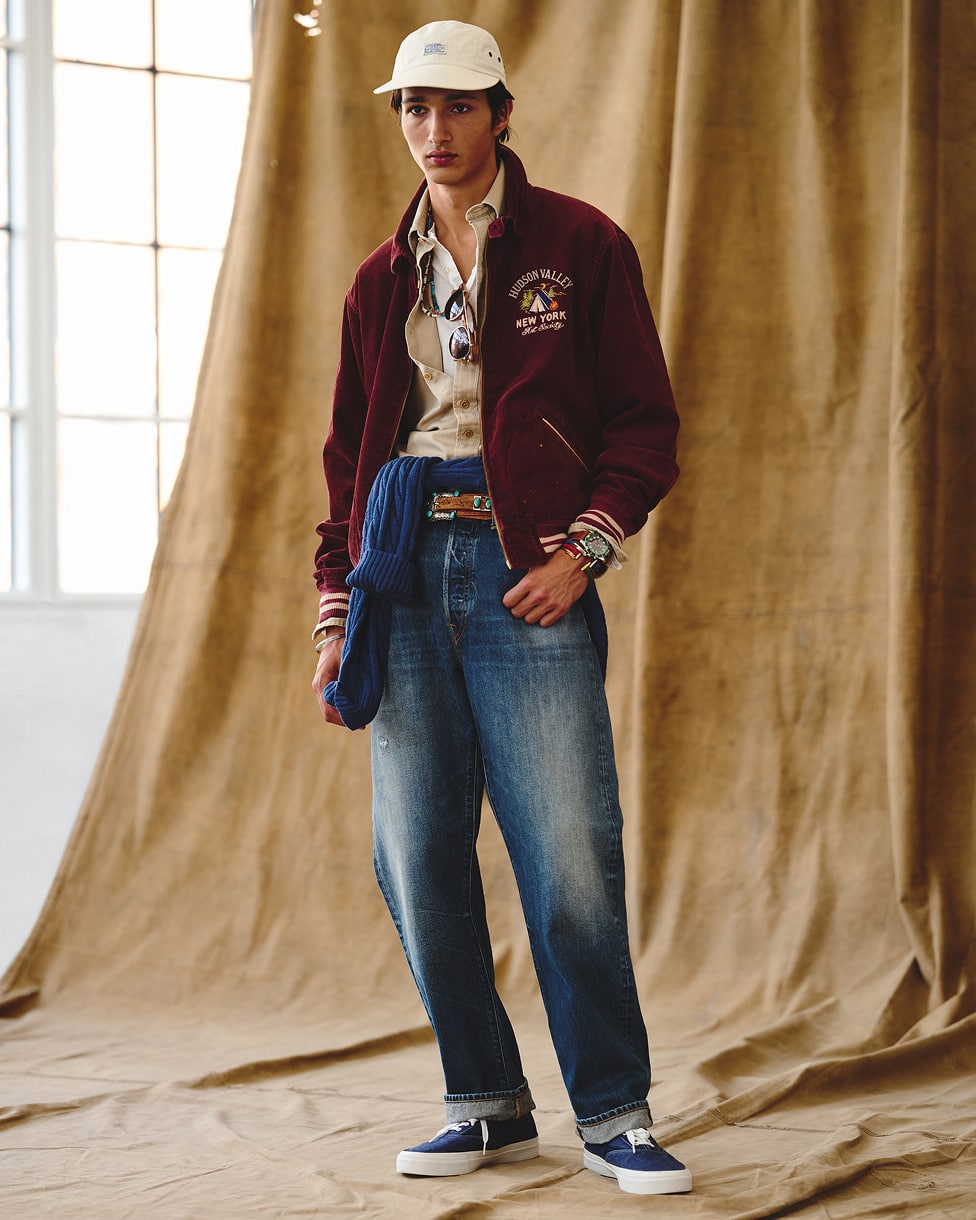 Man wearing a varsity jacket layered over a top and shirt, with jeans and a cap.