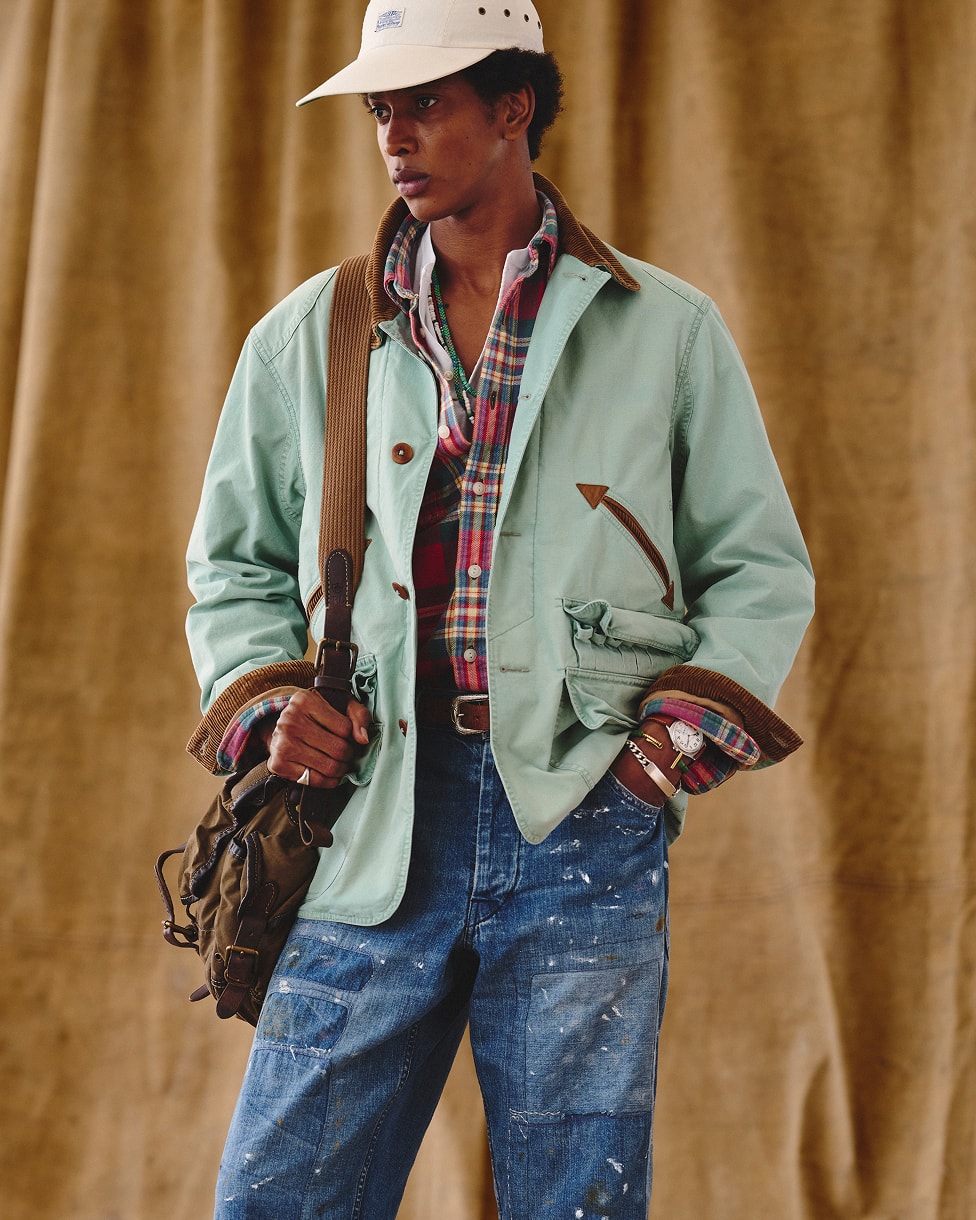 Man wearing a green jacket over a plaid shirt with blue jeans, a cream cap, and a bag.